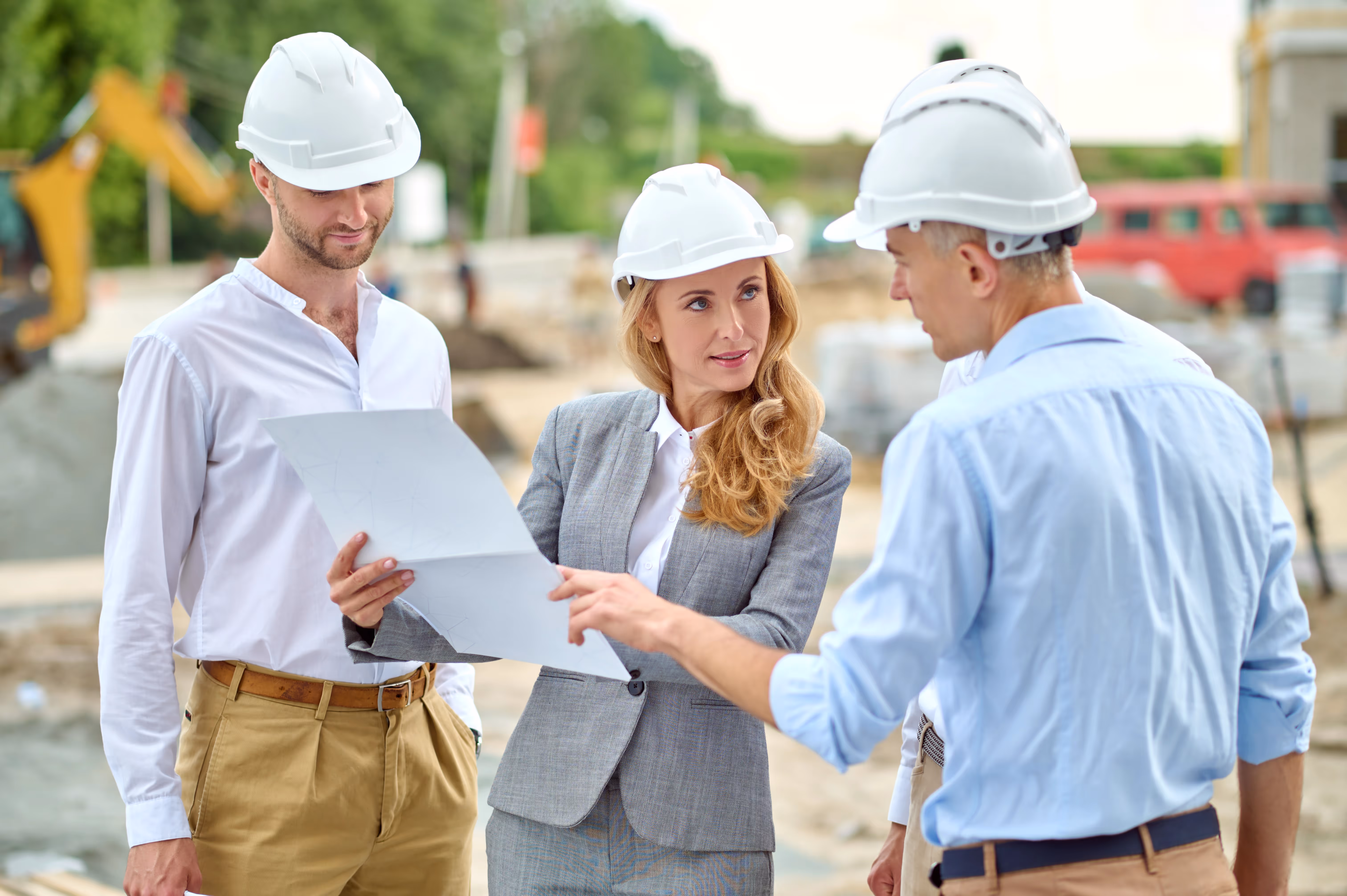 A group of three construction professionals wearing hard hats, discussing project plans at an active construction site. The project owner is holding a document while another is pointing at it, indicating a collaborative discussion.