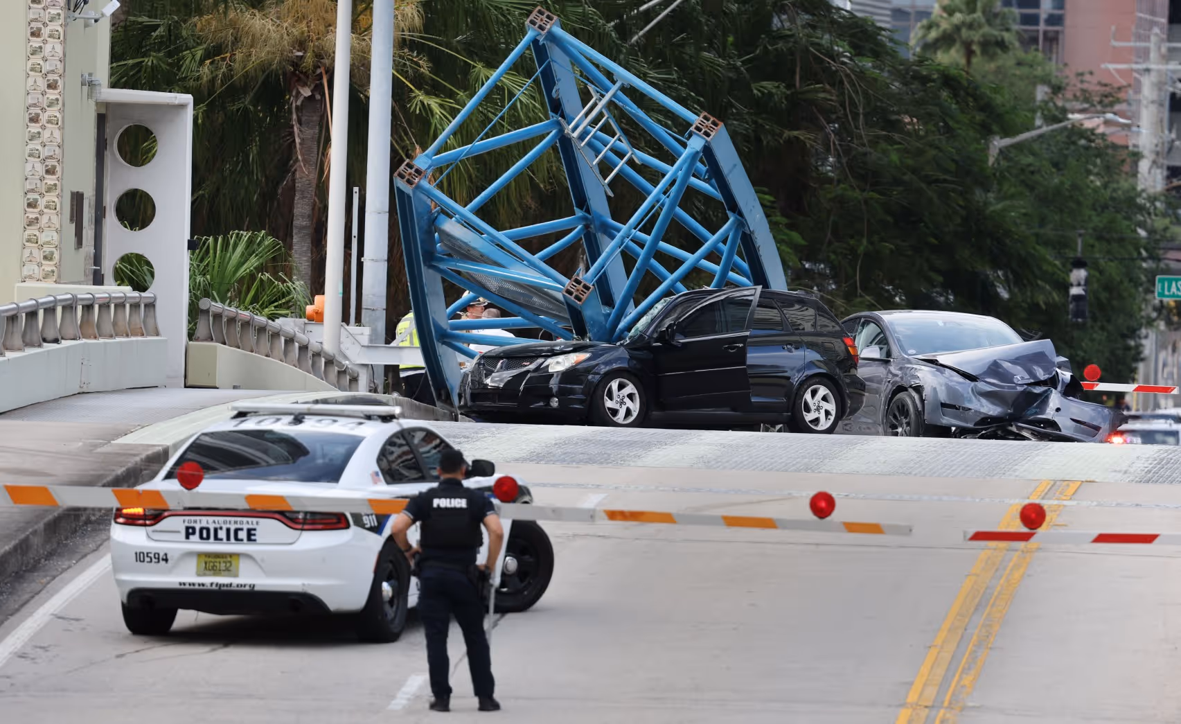 Crumpled cars trapped beneath a large blue crane structure that fell onto the Southeast Third Avenue bridge in Fort Lauderdale, with police and emergency responders at the scene.