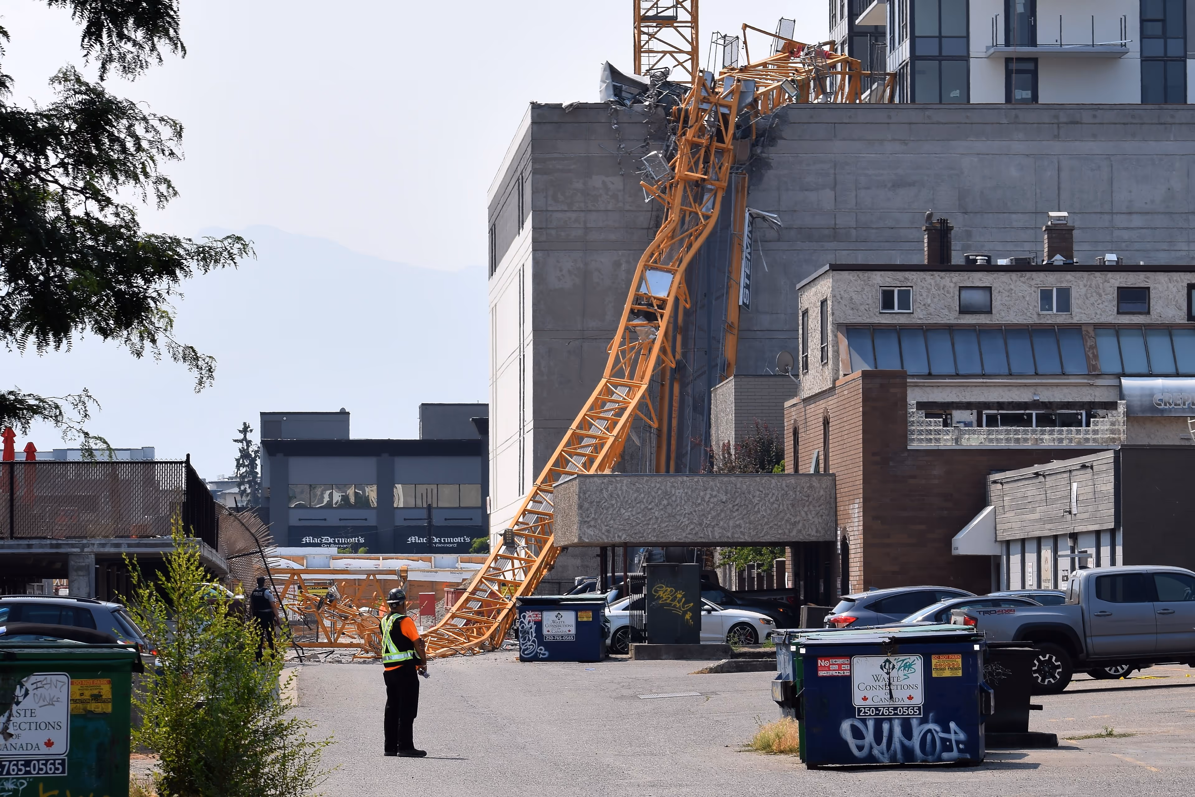 Crumpled yellow crane boom hanging from rooftop and sprawled into alley below, with safety worker standing near debris.