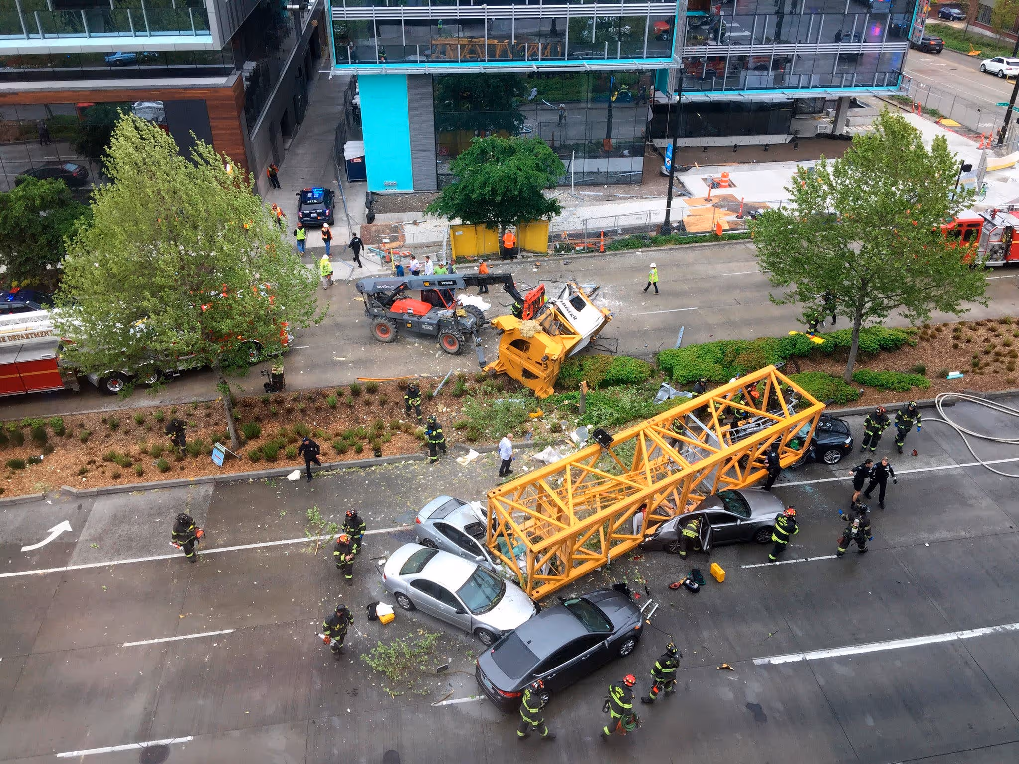 Collapsed yellow crane lying across several crushed cars on a Seattle street, with emergency crews responding at the scene.