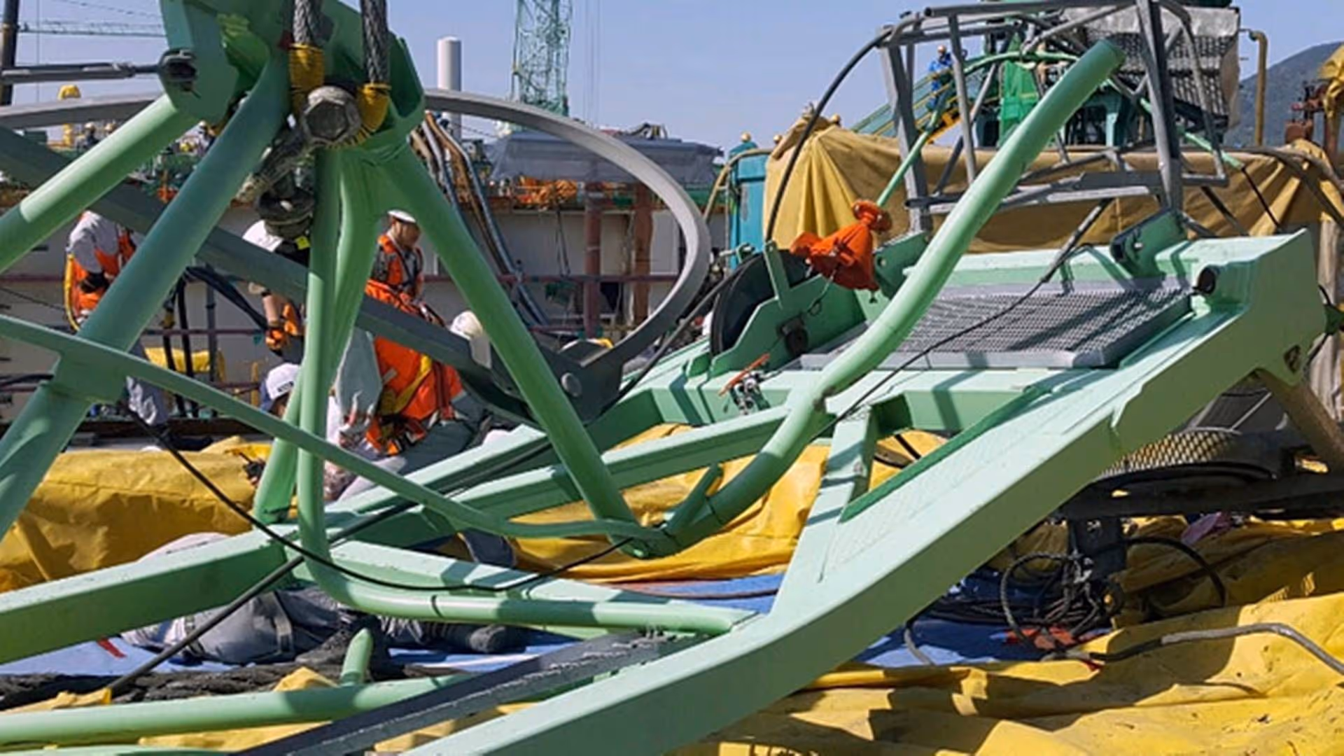 Twisted green crane parts lie across a work site in Geoje, South Korea, as workers in safety gear assess the aftermath of a crane collision.