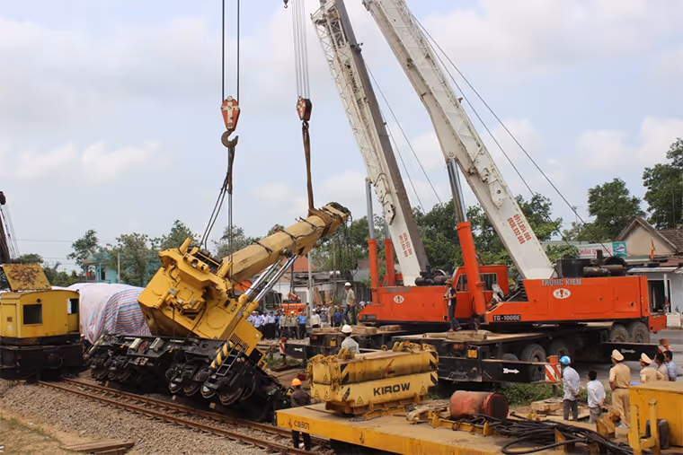 Heavy cranes attempt to lift a toppled yellow gantry crane from railway tracks at Cai Lan Port in Vietnam, with workers and onlookers gathered nearby.