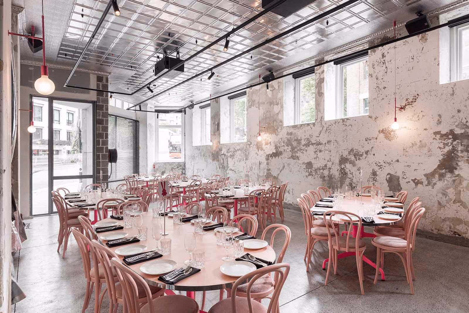 Interior of Chin Chin Sydney restaurant with pink chairs, round tables set for dining, exposed concrete walls, and a polished industrial ceiling.