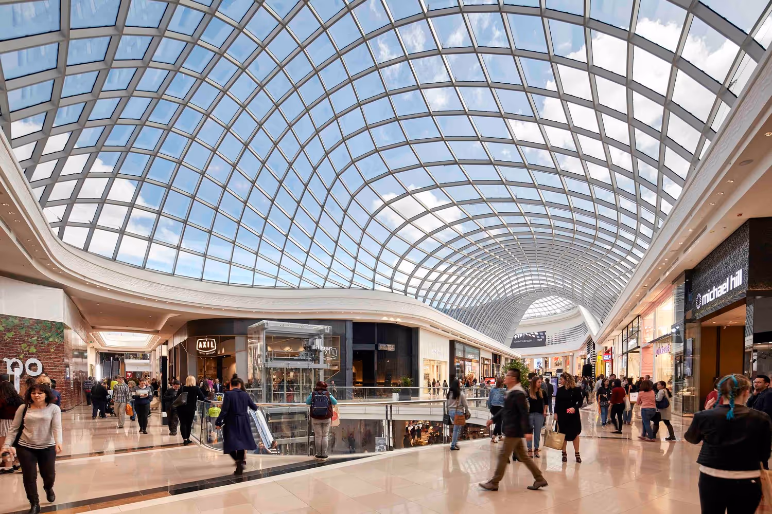 Interior of Chadstone Shopping Centre with curved glass roof, bright natural light, and shoppers walking through high-end retail stores.