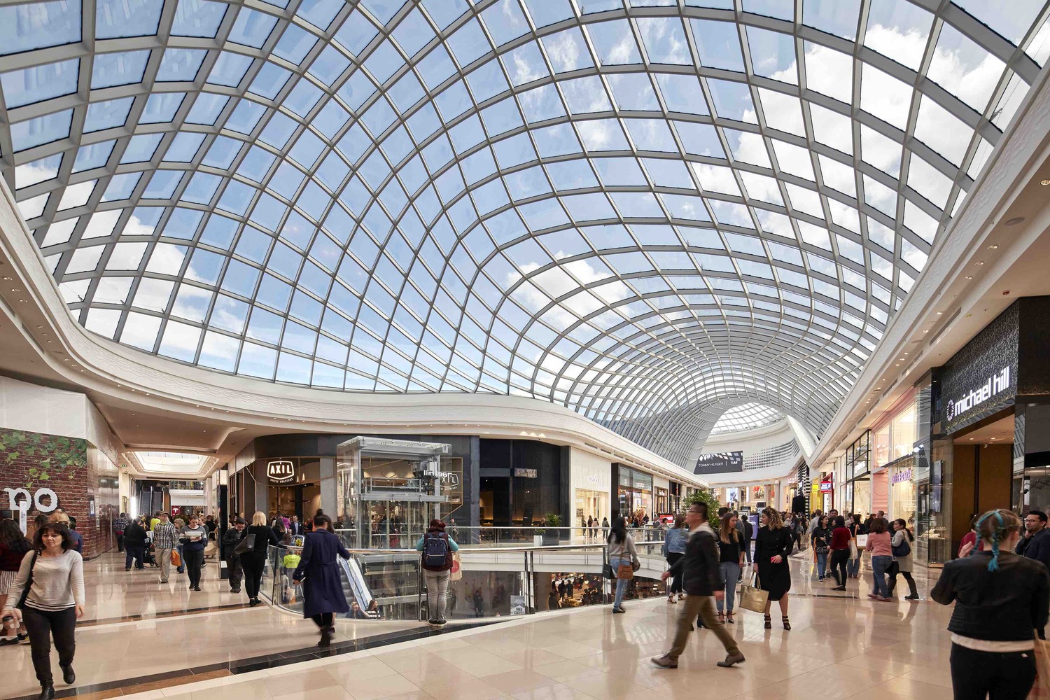 Interior of Chadstone Shopping Centre with curved glass roof, bright natural light, and shoppers walking through high-end retail stores.