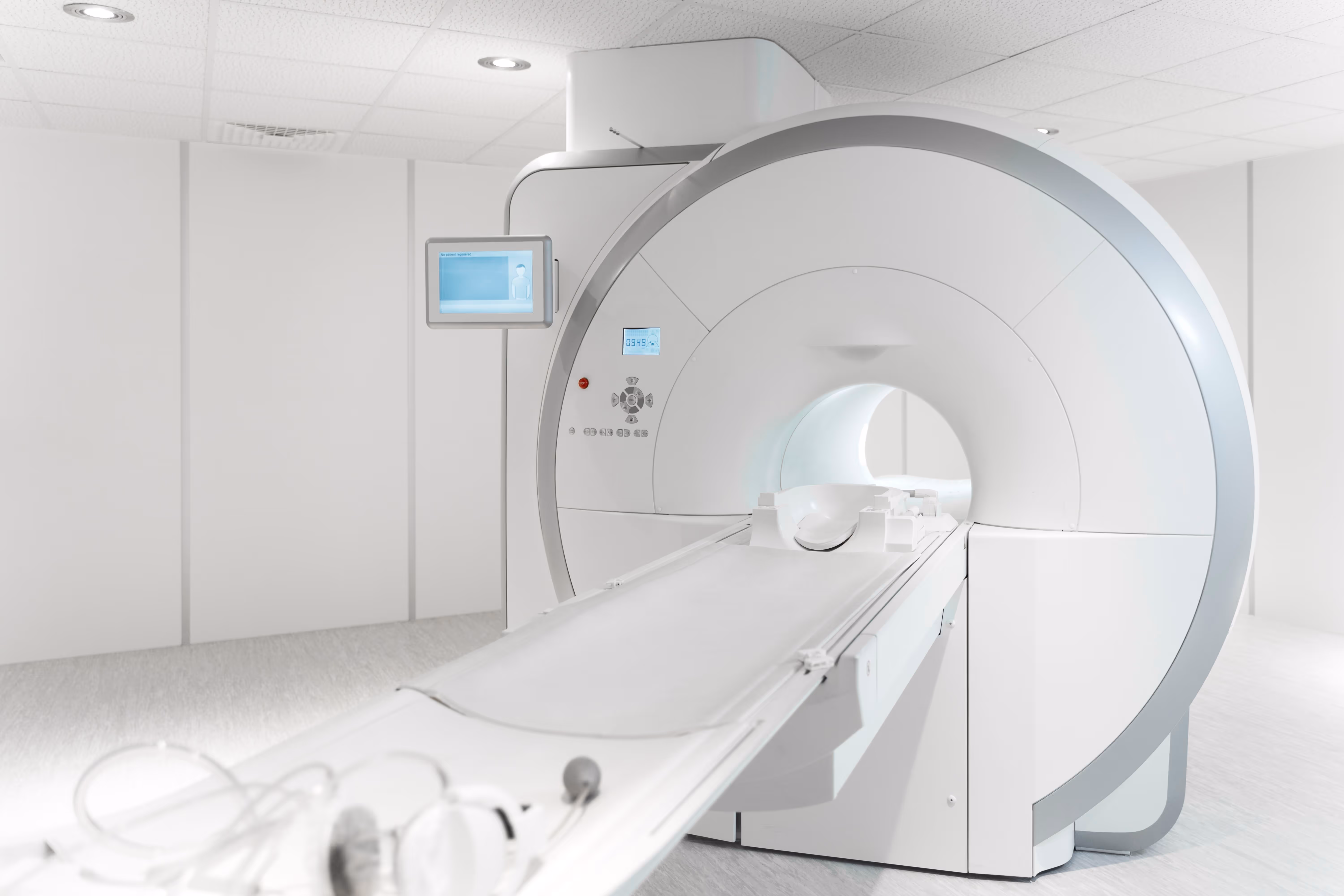 High-angle view of a white CT scanner’s circular gantry and sliding patient table in a bright, sterile examination room with clean walls, ceiling tiles, and an attached control monitor.