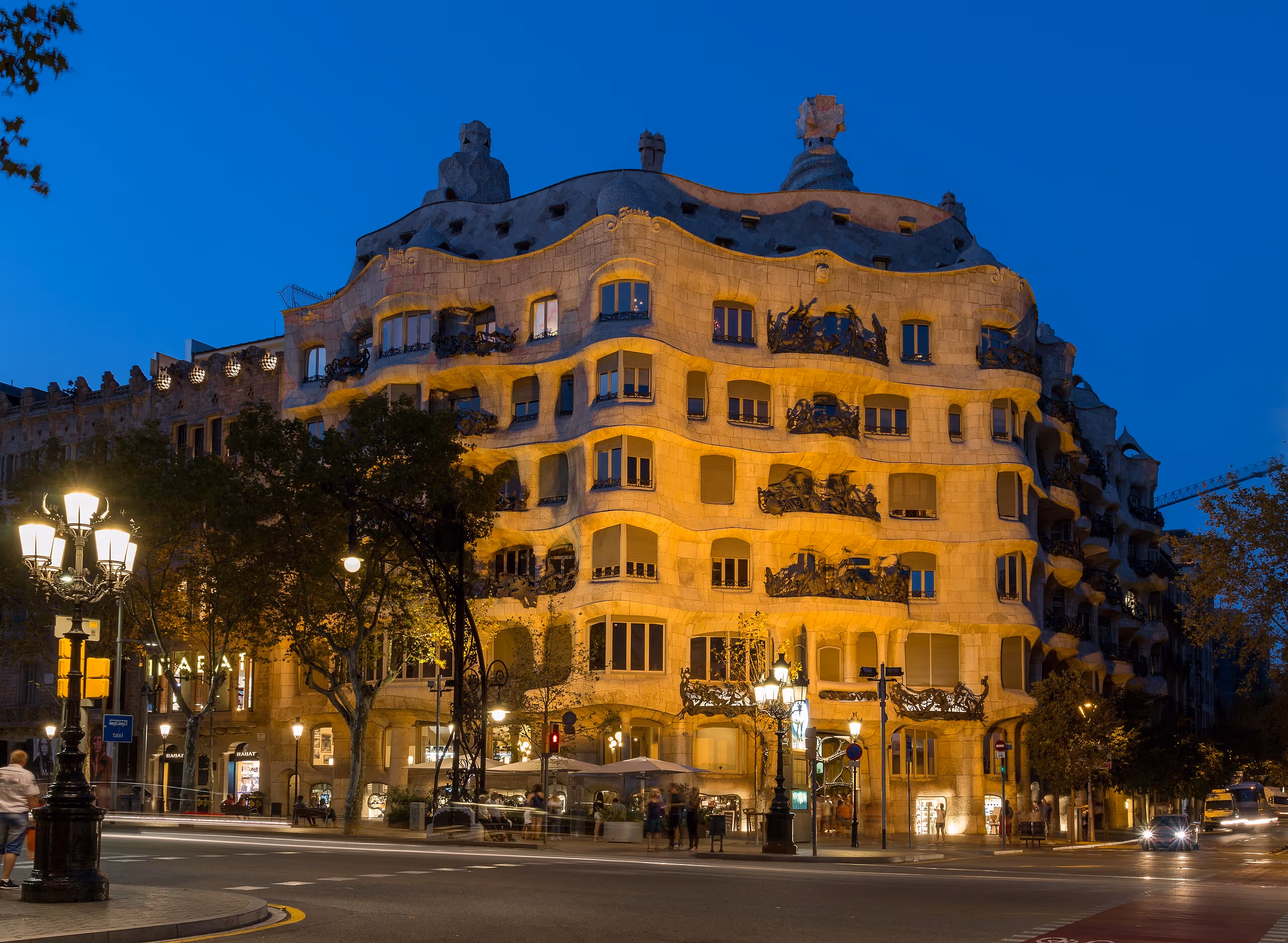 An undulating stone façade with wrought iron balconies and rooftop sculptures, illuminated at dusk.