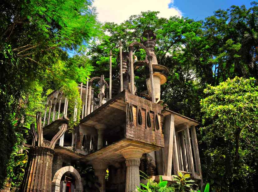 A fantastical sculpture garden filled with towering concrete staircases, columns, and surreal structures set in dense jungle.