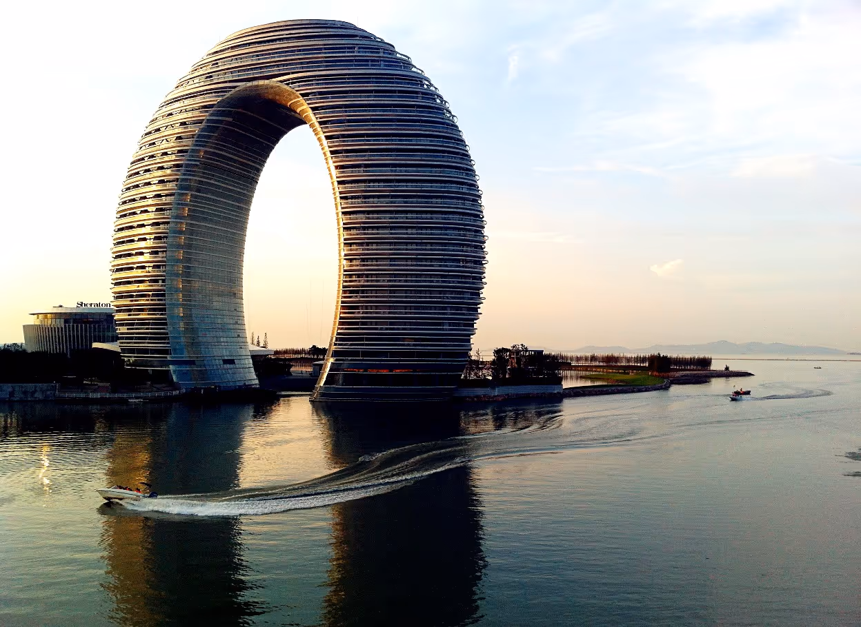 A luxury hotel shaped like a giant ring or doughnut arching over a lake, reflecting in the calm water below.