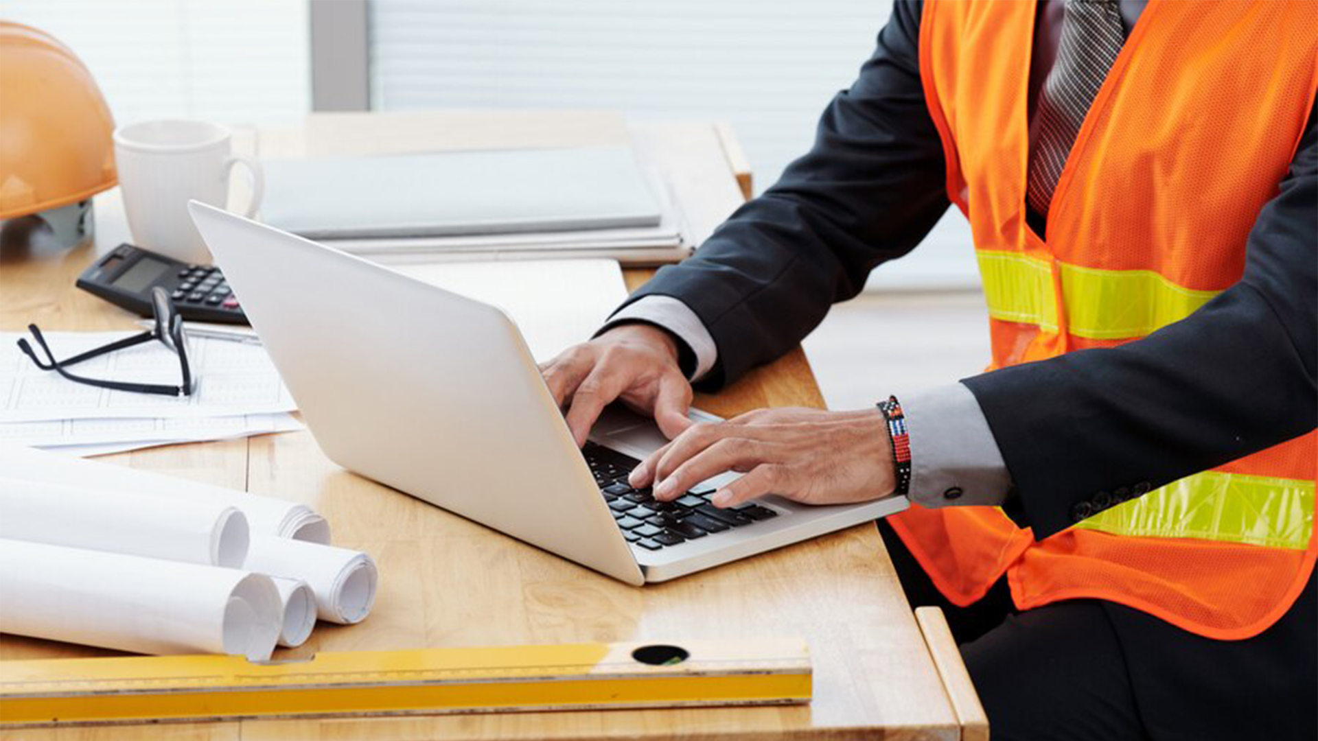 Construction project manager in safety vest working on a laptop at a desk with blueprints, calculator, hard hat, and documents.