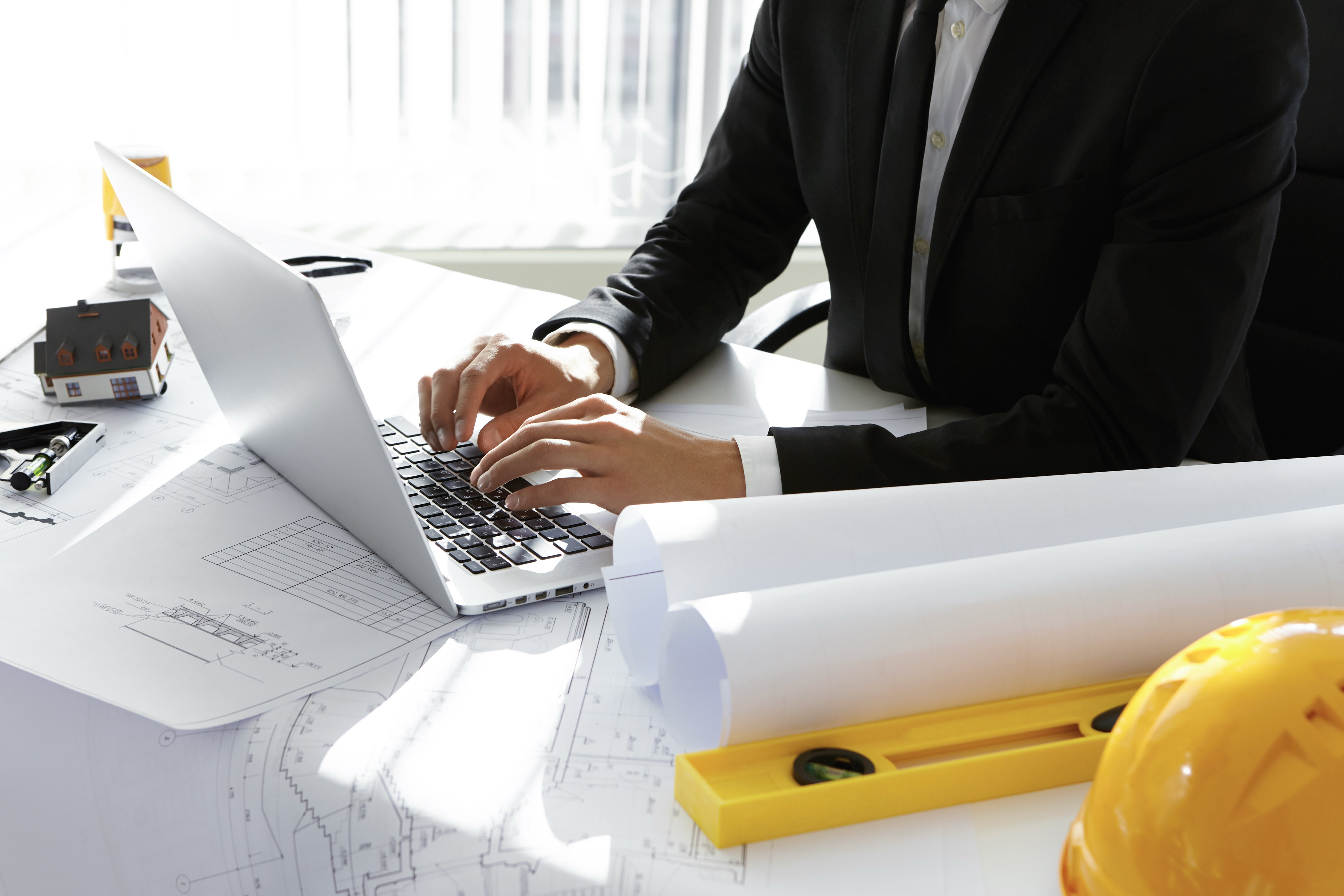 Construction professional reviewing drawings and entering project payment details on a laptop at a desk with plans and safety gear.
