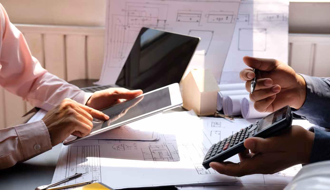 Two construction professionals reviewing job cost data on a tablet and calculator with drawings spread across the table.