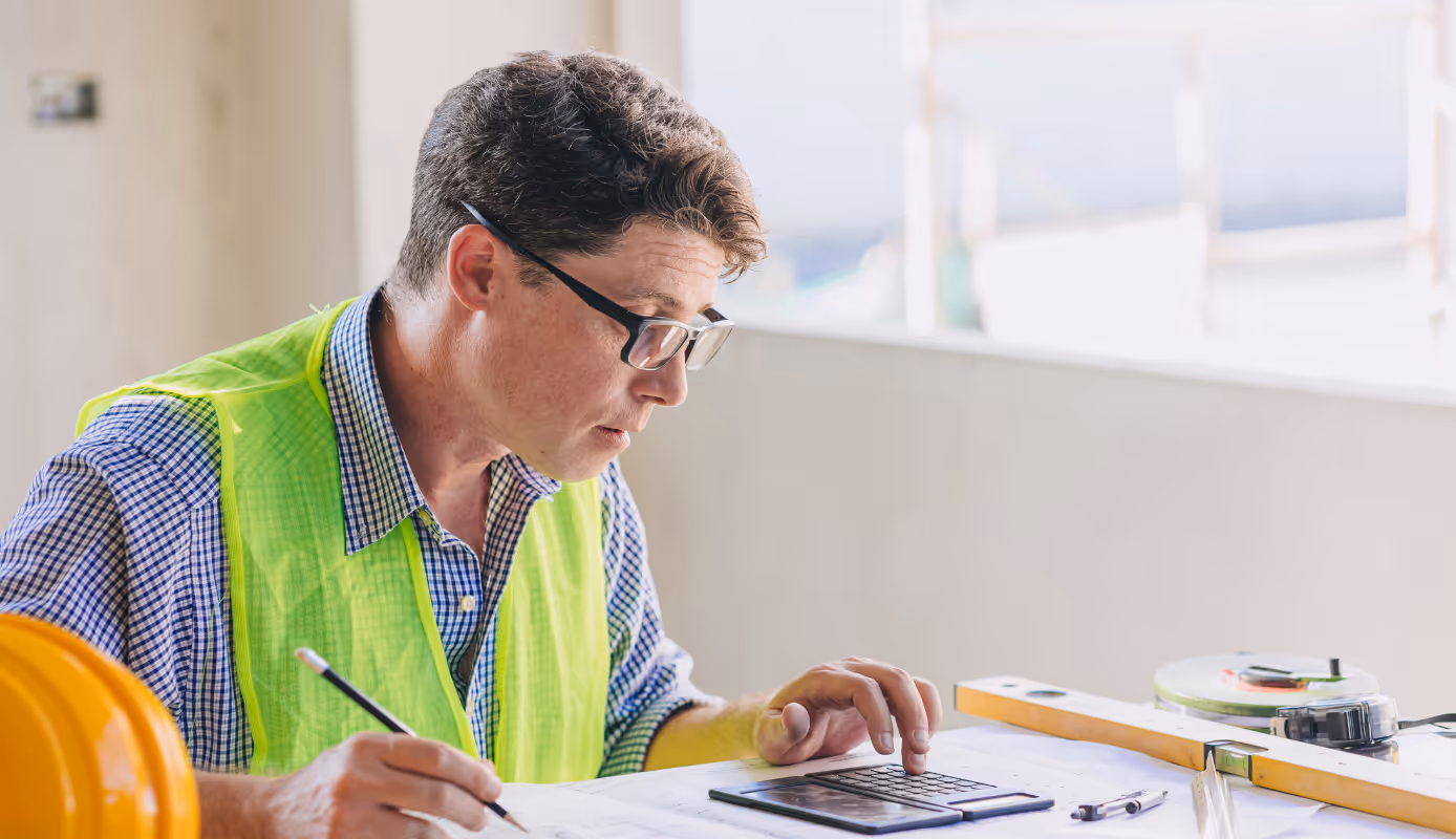 Construction worker in a safety vest calculating job costs on-site using plans and a calculator.