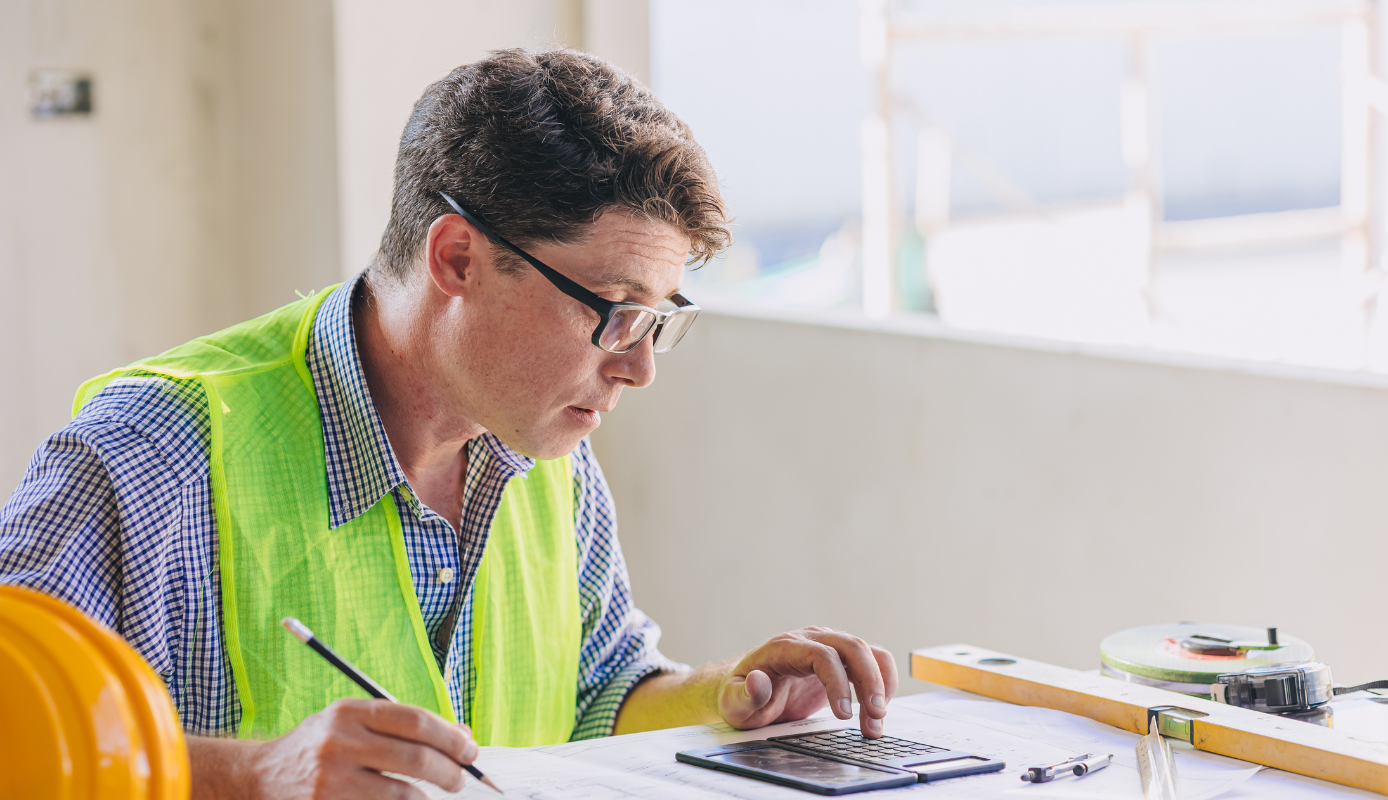 Construction worker in a safety vest calculating job costs on-site using plans and a calculator.