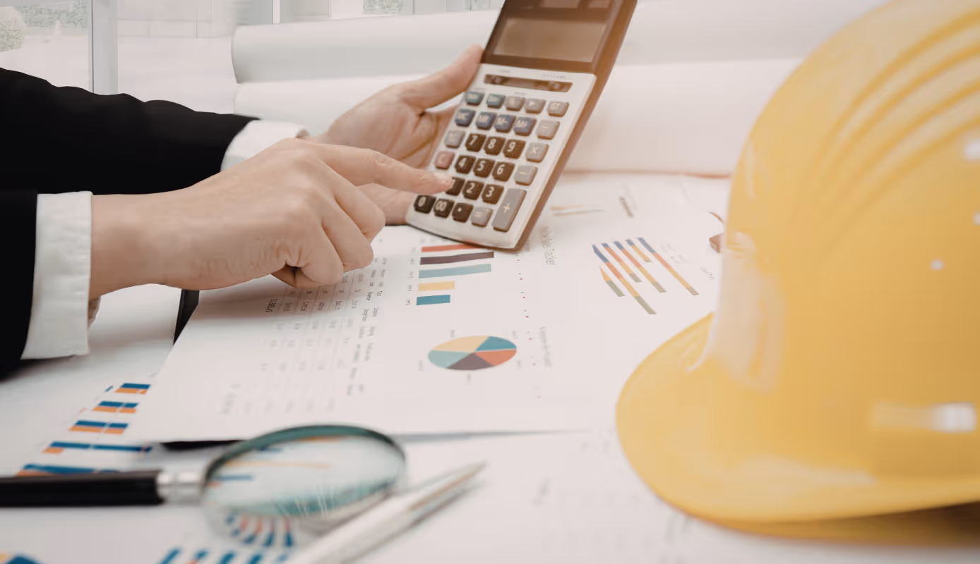 Construction professional using a calculator to review project cost reports and budget charts on a desk with a hard hat.