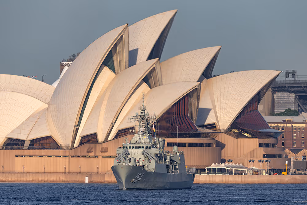 A Royal Australian Navy Anzac-class frigate sails past the Sydney Opera House at sunrise.