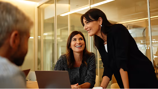 Two program managers collaborating in a modern office meeting room. One is seated with a laptop, smiling, while the other stands confidently, engaging the team in discussion. A third colleague is partially visible in the foreground, emphasizing coordination, leadership, and cross-project collaboration.