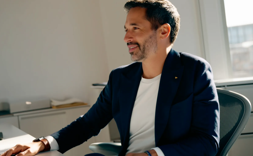 A project executive wearing a navy blazer and white shirt sits at a desk in a modern, sunlit office. He is smiling and looking to the side, appearing engaged and confident. A minimal, professional setting with natural light enhances the corporate atmosphere.