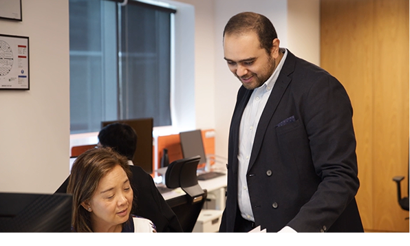 A project manager in a dark blazer stands beside a seated colleague in an office, smiling as they discuss a document. The environment features desks, monitors, and a collaborative team setting, suggesting a professional and engaged workplace atmosphere.