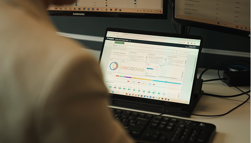 A project manager works at a desk with a laptop displaying the Mastt project management platform. The screen shows dashboards with project status indicators, timelines, and data widgets. Additional monitors are visible in the background, reflecting a data-driven and tech-enabled work environment