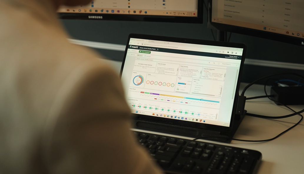 A person is viewing the Mastt AI-powered construction project management dashboard on a laptop screen. The dashboard displays project performance metrics, visual charts, progress indicators, and a timeline. The workspace is equipped with multiple monitors, reflecting a data-driven, professional project control environment