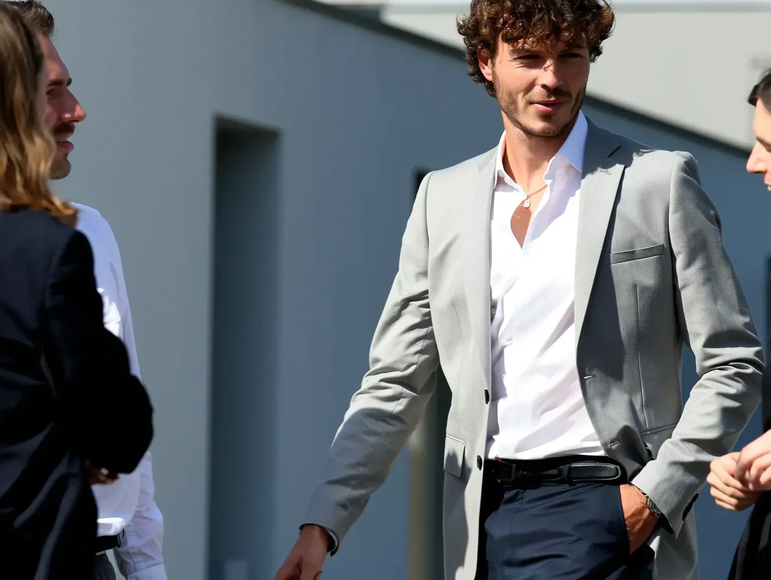 A project manager in a light grey blazer and white shirt stands outdoors in conversation with colleagues. He has one hand in his pocket and a confident expression, engaging in a casual yet professional discussion in a modern building setting.