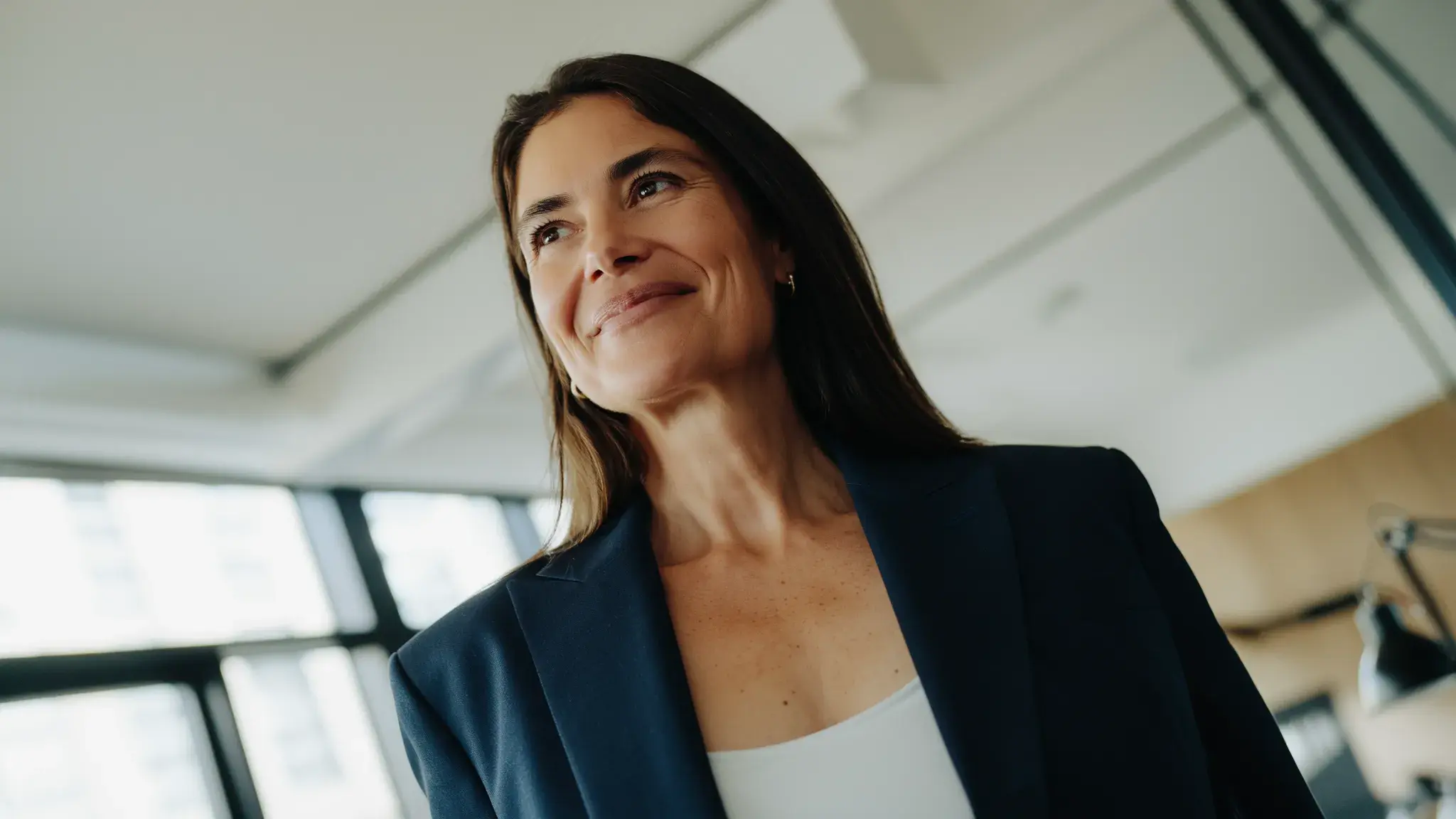 A confident program manager wearing a navy blazer and white top smiles softly while standing in a modern office with large windows. The natural lighting and contemporary workspace convey a professional and forward-thinking environment