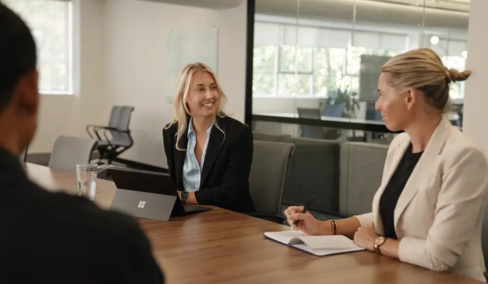 Two project managers in business attire are engaged in a meeting around a conference table in a modern office. One is smiling while using a laptop, and the other is taking notes in a notebook. Two colleagues are partially visible in the foreground, indicating a collaborative team setting.