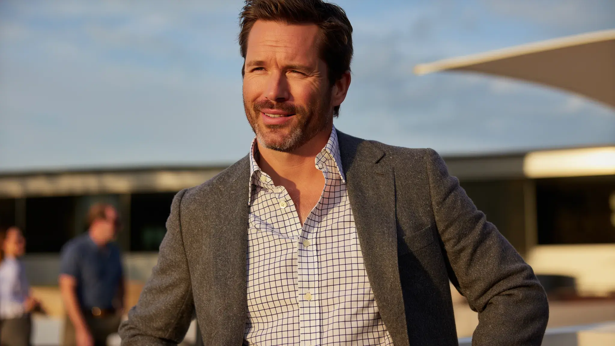 An owner's representative in construction stands outdoors during golden hour, wearing a grey blazer and a checkered dress shirt. He appears confident and engaged, with a slight smile, while colleagues converse in the background on a construction or development site.