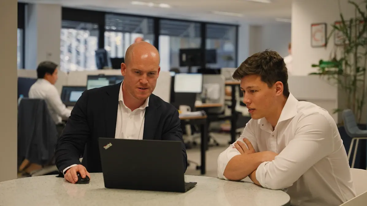 Two development managers in business attire collaborate at a round table in a modern office. One is operating a laptop while the other listens attentively with arms folded. The background shows a busy workspace with colleagues working at desks and a professional, tech-driven atmosphere.