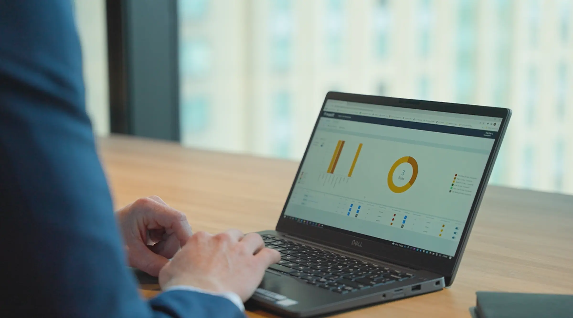 A person in a blue blazer works on a laptop displaying a Mastt risk dashboard interface. The screen shows visual data including bar charts and a donut chart summarizing risk categories and quantities. The workspace is set near large windows overlooking a cityscape, creating a professional and focused environment for project risk analysis.
