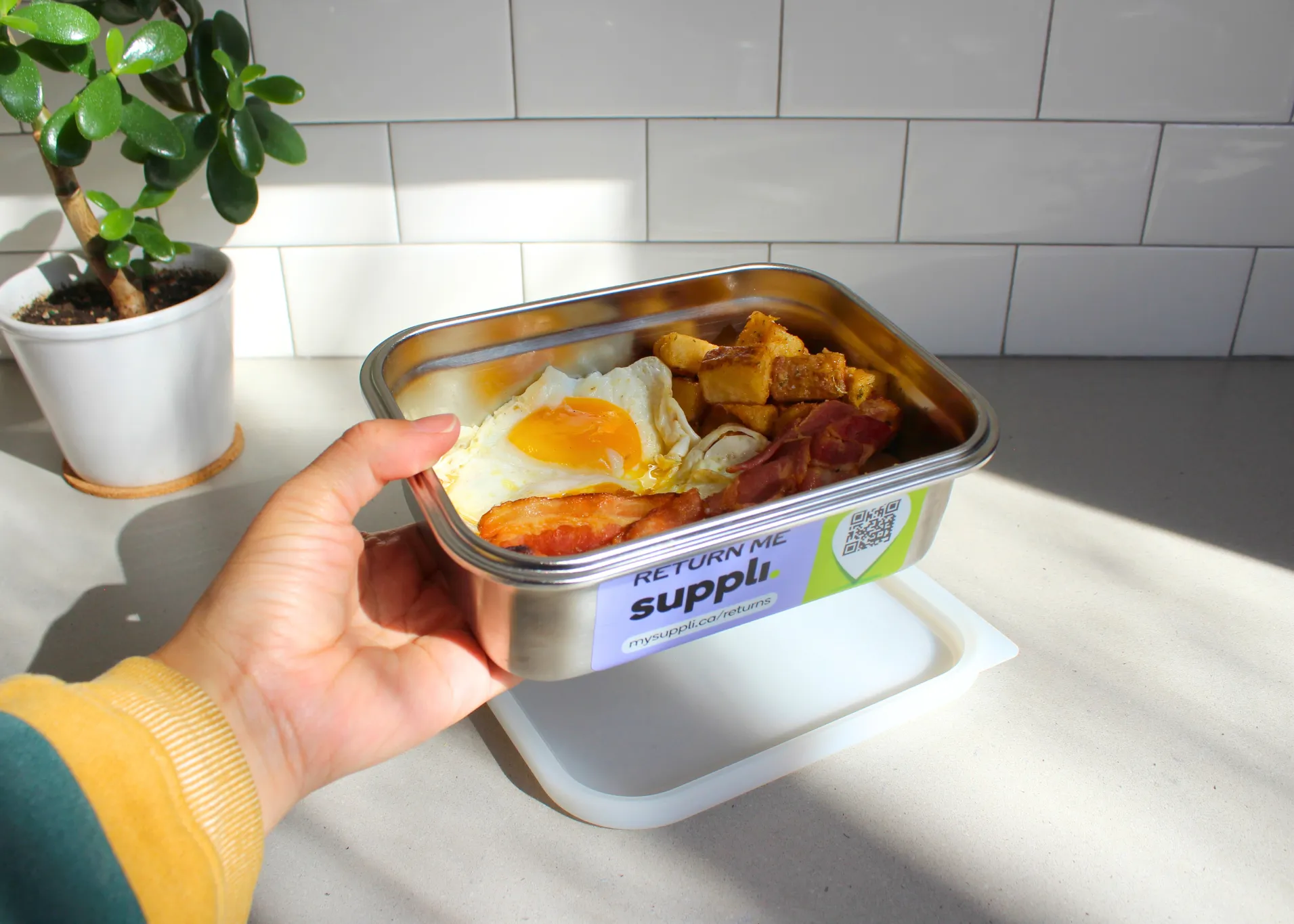Hand holding a stainless steel food container with cooked breakfast items including a fried egg, bacon, and cubed potatoes, with a label reading 'RETURN ME suppli.' and a QR code on the side, against a kitchen countertop with white subway tile backsplash and a small potted plant.