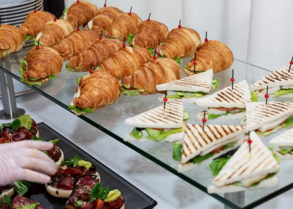 Tray of assorted sandwiches including croissant sandwiches with lettuce and triangular toasted sandwiches with grill marks, each held with a toothpick with a red bead, arranged on a glass table.