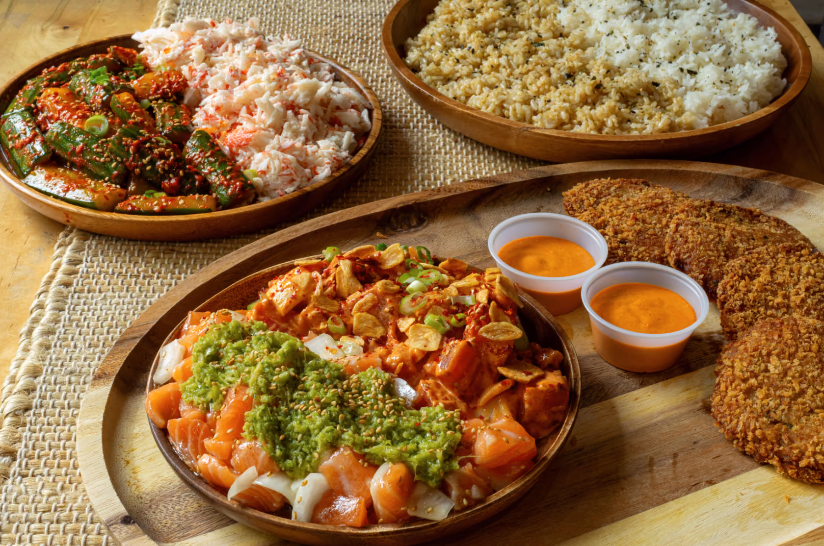 Wooden plates with diverse food: a bowl of rice and seasoned rice, a plate with cucumber kimchi and shredded crab meat, a platter with crispy patties served with two cups of orange dipping sauce, and a bowl containing diced raw salmon topped with green sauce and garlic chips.