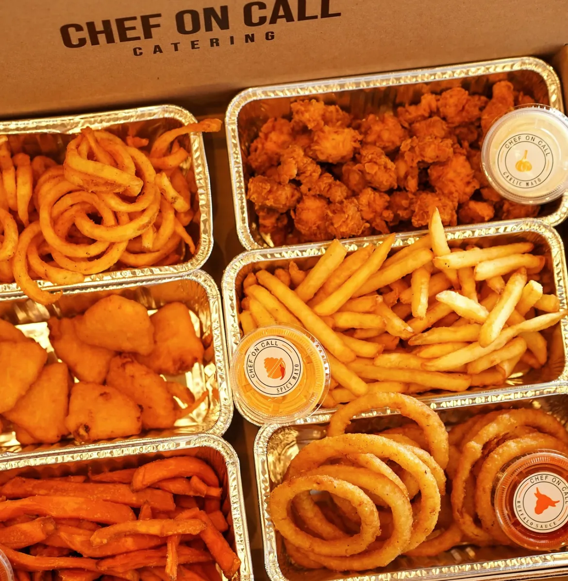 Various fried foods in aluminum trays including curly fries, popcorn chicken, regular fries with spicy mayo sauce, chicken nuggets, sweet potato fries, and onion rings with buffalo sauce, inside a box labeled Chef On Call Catering.