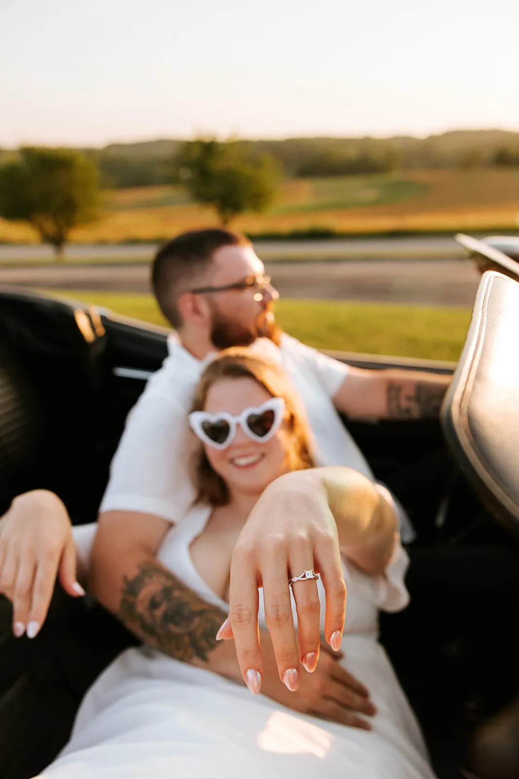 Woman wearing heart-shaped sunglasses showing an engagement ring on her hand while sitting in a car with a man behind her.