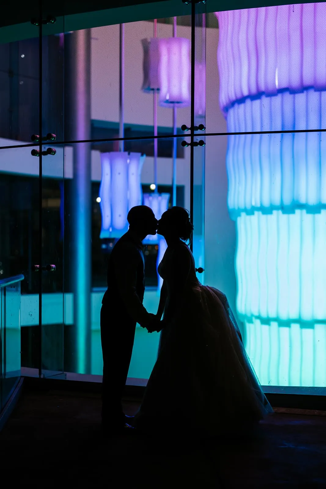 Silhouette of a couple kissing and holding hands in front of a glass wall with blue and purple illuminated modern light fixtures.