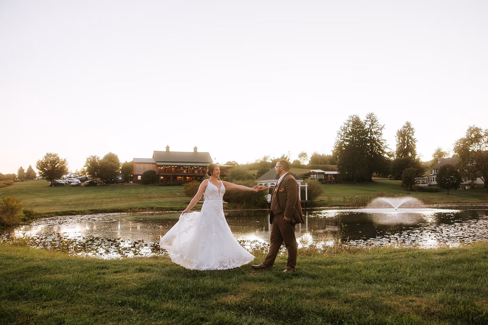 Bride in a white wedding dress and groom in a brown suit dancing near a pond with a fountain and a barn in the background at sunset.