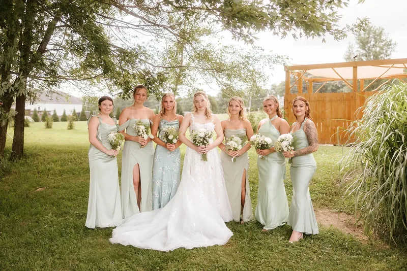 Bride and bridesmaids in sage dresses holding bouquets outdoors.