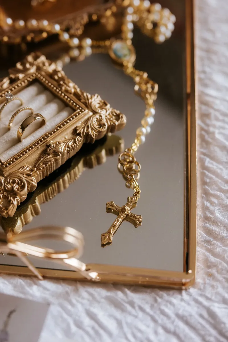 Wedding jewelry and a gold rosary arranged on a mirrored tray.
