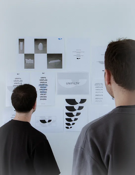 Deux hommes vus de dos regardant un mur blanc avec plusieurs affiches imprimées contenant des textes et des formes graphiques noires.