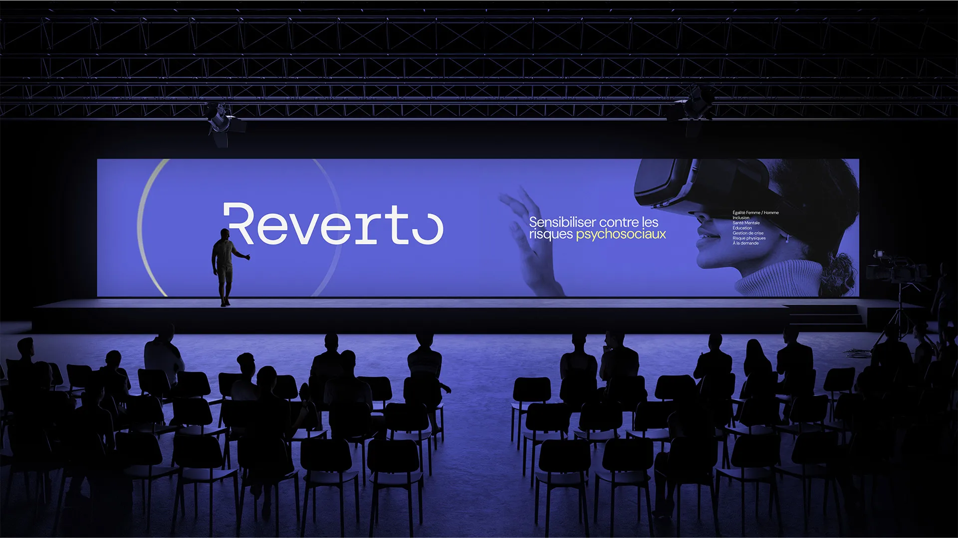 Une silhouette présente sur scène devant un écran bleu affichant le texte "Reverto" et "Sensibiliser contre les risques psychosociaux" avec l'image d'une personne portant un casque de réalité virtuelle.