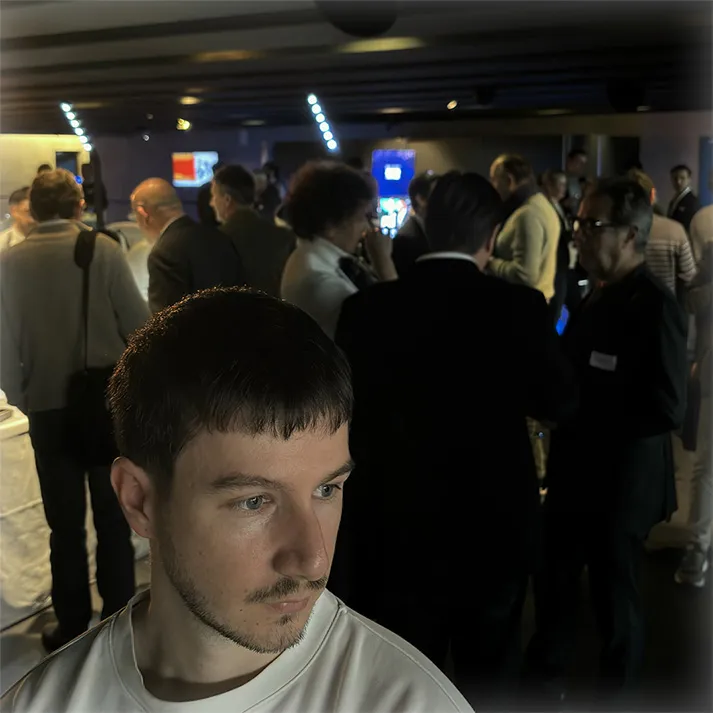 Un homme avec des cheveux courts et une barbe naissante regarde vers la gauche, avec une foule floue en arrière-plan lors d'un événement en intérieur.