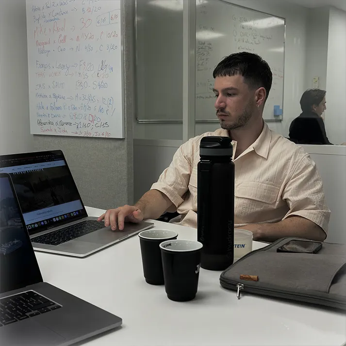 Homme assis à une table de travail, utilisant un ordinateur portable avec une grande bouteille noire et plusieurs gobelets devant lui.