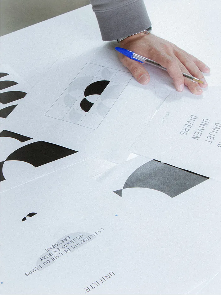 Main tenant un stylo bleu au-dessus de feuilles avec des dessins et textes en français sur une table blanche.