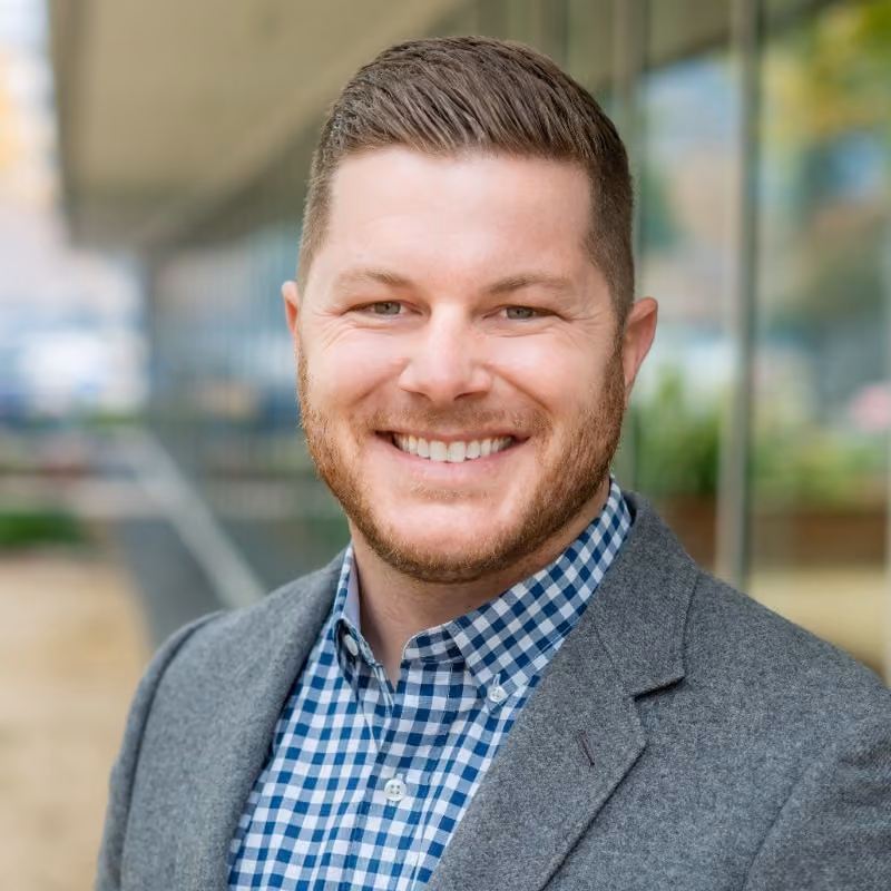 Smiling man with short hair and beard wearing a gray blazer and blue checkered shirt.