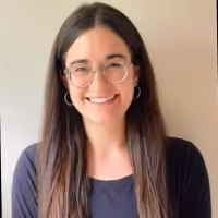 Smiling woman with long brown hair, glasses, and hoop earrings against a plain background.