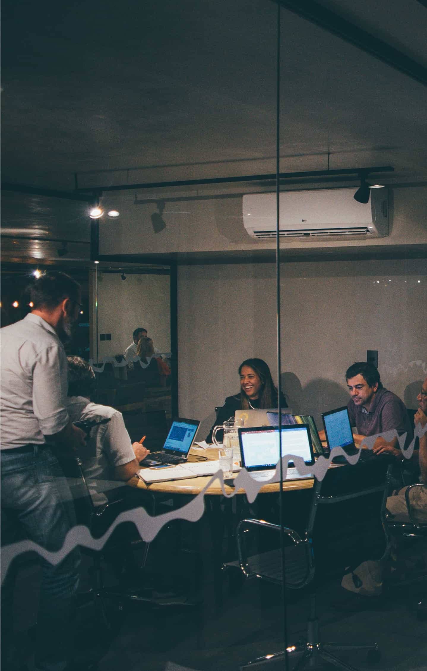 Team collaborating in a glass-walled meeting room, working on laptops and documents.