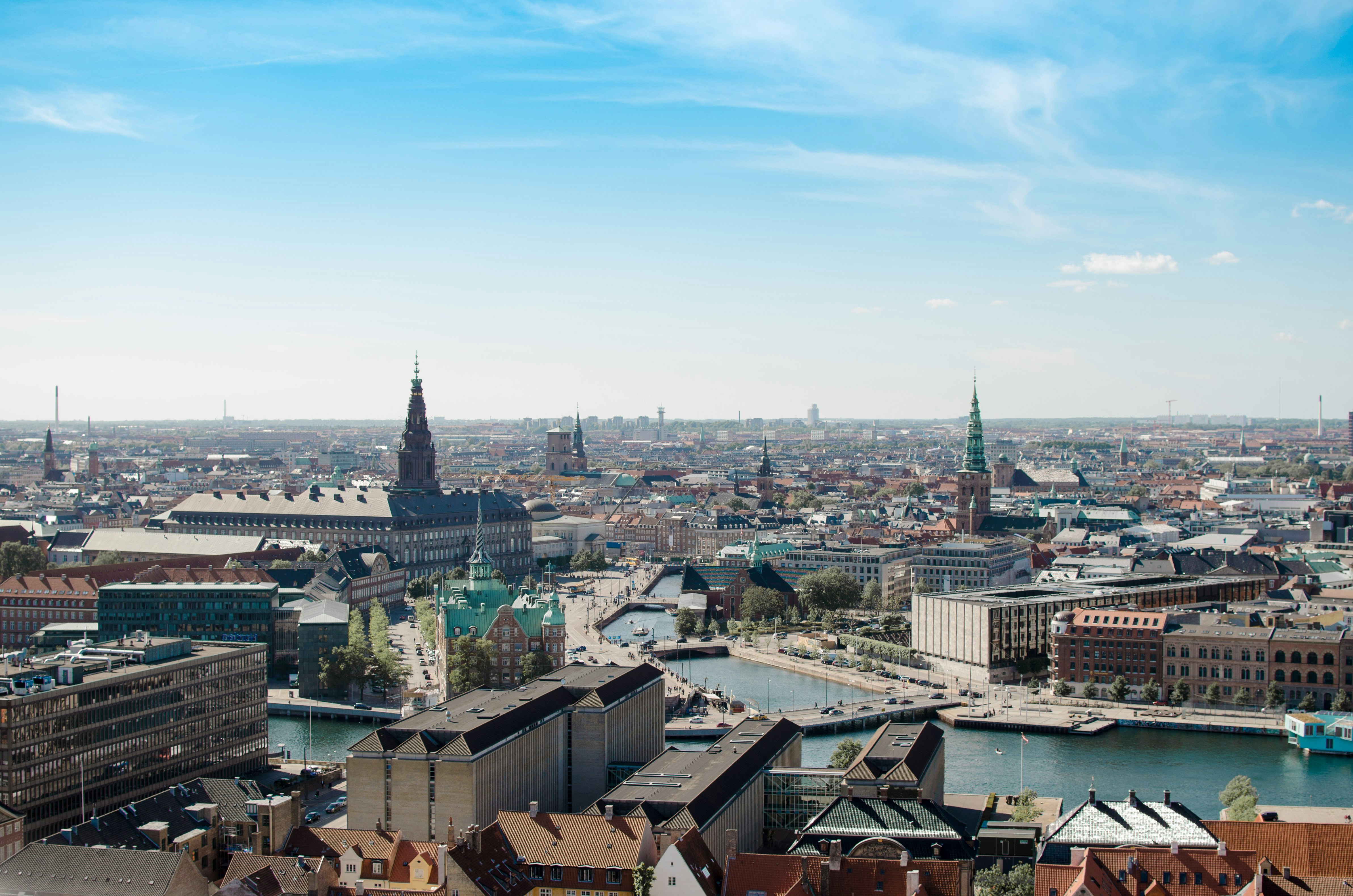 Aerial view of Copenhagen, Denmark, featuring historic buildings, church spires, canals, and modern architecture under a clear blue sky.