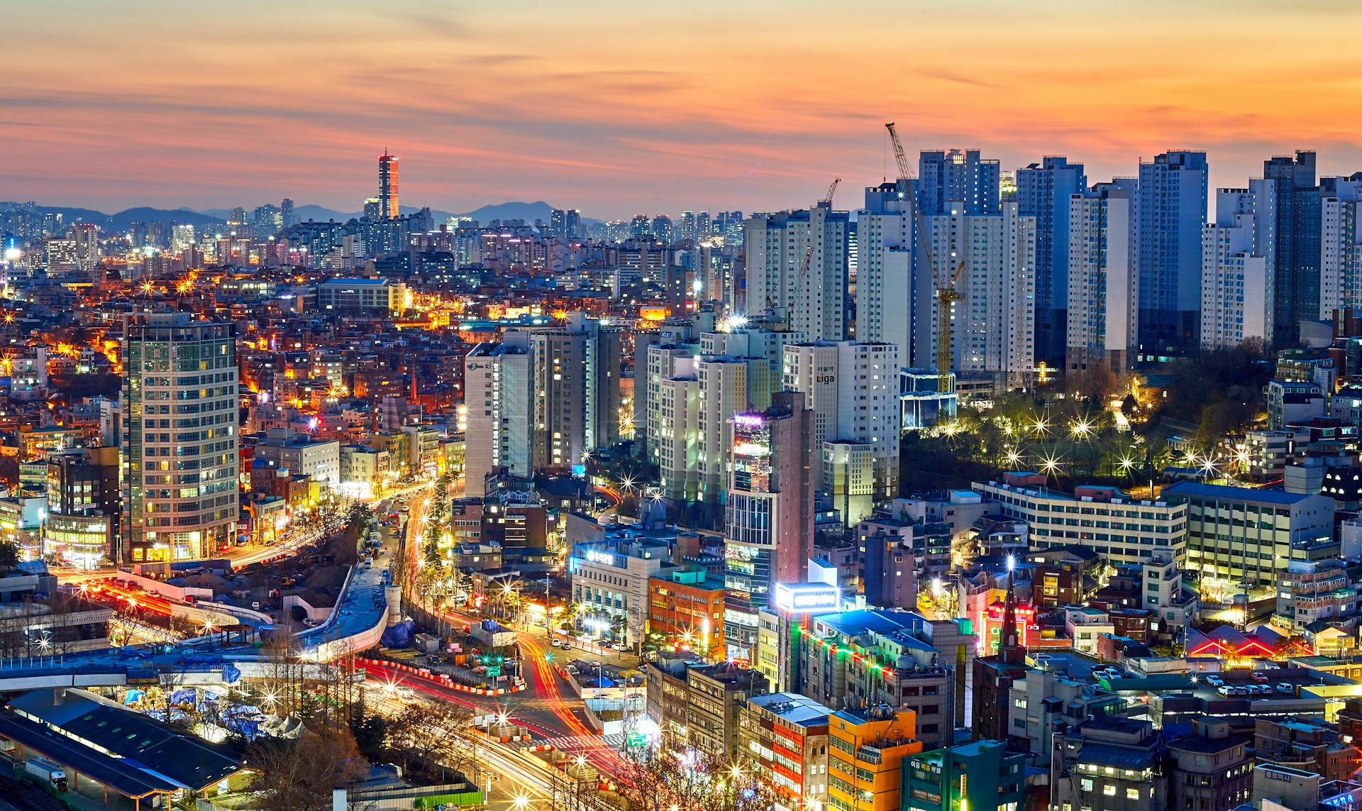 Vibrant cityscape of Seoul, South Korea at sunset, featuring illuminated high-rise buildings, busy roads with light trails, and a colorful urban skyline.